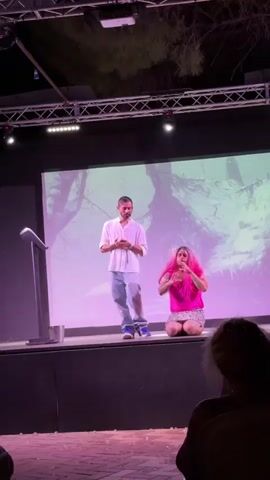 The image depicts a performance on a stage with a large screen in the background displaying an abstract, possibly natural, image in shades of purple and white. Two individuals are on stage. On the left, a man in a white shirt and light-colored pants stands at a podium, appearing to speak or present. On the right, a person with vibrant pink hair, wearing a pink top and a patterned skirt, kneels on the stage. They are making expressive hand gestures and seem to be engaged in an emotional performance. The stage is lit with purple and white lights, casting a theatrical atmosphere. In the foreground, scattered transparent pieces, resembling broken glass or confetti, lie on the floor. The scene appears to be taking place outdoors at night, evidenced by the dark sky and the stage lighting. The presence of trees visible above the stage structure further suggests an outdoor venue. The audience is partially visible in the bottom right corner, their backs to the camera. The overall mood is dramatic and engaging.