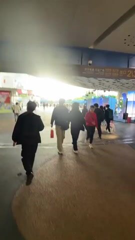 The video shows several people walking through a covered outdoor walkway. There is a couple holding hands, a young person running, and other pedestrians strolling in both directions. Buildings are visible in the distance, and signs hang above the walkway. The area is well-lit, and the ground appears to be a mix of concrete and pavement.