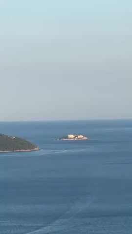 The video shows a wide view of a body of water, likely the sea, with a clear sky above. In the distance, there are two landmasses. The larger landmass on the left appears to be a hilly, tree-covered island or coastline. Closer to the center of the frame, a smaller island with a man-made structure on it is visible. The structure appears to be light-colored and rectangular, possibly a building or a complex of buildings. The water is a deep blue and appears calm, with some faint boat wakes creating linear patterns on the surface. A large white cruise ship is seen on the left side of the frame, moving away from the coastline. Smaller boats are also visible, creating trails on the water. The lighting suggests it is daytime, likely late afternoon or early morning due to the soft light.