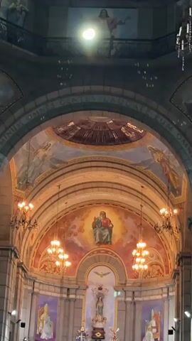 The video shows the interior of a grand church. The camera is angled upwards, focusing on a large, ornate altar area. Above the altar, there are several murals depicting religious figures. To the left and right of the altar are large chandeliers with multiple lights. In the center, above a statue of a religious figure, is a mural of a dove. The overall lighting is dim, with the chandeliers and some spotlights providing illumination. The footage appears to be of low quality, with some blurring and digital noise. The scene is static, with no movement visible. There are no people clearly visible in the frame.