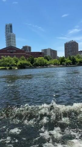 The video begins with a low-angle, wide shot of a body of water, likely a river, with a foamy wake from a moving boat in the foreground. The water is dark blue and choppy, reflecting the bright blue sky and scattered white clouds above. Beyond the water, a tree-lined bank is visible, with lush green trees obscuring some of the background. Behind the trees, a cityscape emerges, featuring several multi-story buildings. The buildings vary in architectural style and height, including a tall, modern skyscraper with white vertical accents, several brick apartment buildings, and a large, dark brick building. The overall scene suggests a sunny day in an urban environment, viewed from a boat on the water. The timestamp indicates the video is approximately 4 seconds long, with the text "It is a gorgeous day here on the Charles" appearing at the end.