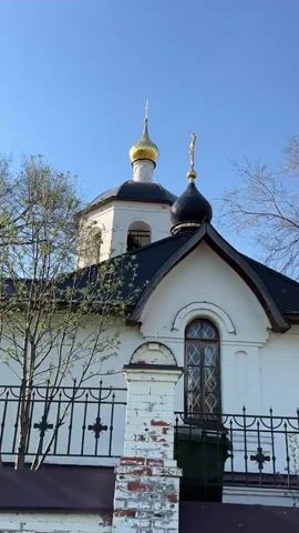 The video shows an exterior shot of a white building with an Orthodox-style dome. The building has a black roof and a white brick exterior. There are windows with intricate designs. A black metal fence adorned with cross symbols runs along the front of the building.  A golden dome rises prominently above the building. Bare trees are visible in the background against a clear blue sky. The shot pans to provide a broader view of the building's architecture, including a rounded annex. The overall setting appears to be a sunny day in an outdoor location with historical or religious significance.