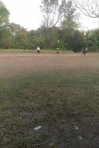 A group of people are playing soccer on a dirt field. The field is surrounded by trees and vegetation. There is a goalpost on the left side of the frame. The sky is clear and there is sunlight.