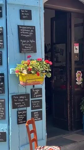 The video shows the exterior of a cafe or restaurant with people seated at tables outside. The establishment has blue shutters on either side of the entrance. The entrance itself is dark, with a sign above it. There are chalkboards with writing on the shutters and walls. Several potted plants are visible, including one with red and pink flowers. A person wearing a white hat and white clothing is seated on a chair to the right of the entrance, holding a drink. Further down the street, more tables and umbrellas are set up, with several groups of people dining and socializing. A man wearing a dark t-shirt and jeans is standing near the tables on the right, possibly a waiter or staff member. The overall setting appears to be a sunny day in a European-style town or village.
