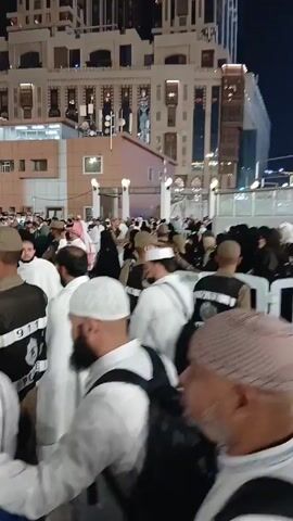 The video shows a large crowd of people in a plaza at night. Many are wearing traditional Islamic clothing, like white thobes and head coverings. Some people wear civilian clothes. There are individuals wearing brown uniforms with "POLICE" printed on their vests. They stand interspersed within the crowd, maintaining order. The environment includes tall buildings, one appearing to be under construction with exposed scaffolding and lit by string lights. The crowd is dense but moving slowly, suggesting controlled movement or waiting.