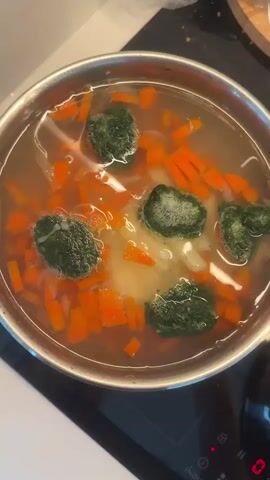 In a kitchen setting, a person's hand uses a black spoon to stir a metal pot filled with soup on an induction cooktop. The soup contains visible ingredients such as diced orange carrots, lighter-colored vegetables (possibly potatoes or onions), and several clumps of a dark green vegetable, likely spinach or broccoli. The liquid in the pot is clear broth or water, actively bubbling. The induction cooktop displays "INDUCTION" on its control panel. The scene takes place in Savenki, Russia.