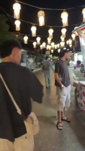 Two young men are walking down a street at night. There are streetlights in the shape of lanterns overhead. One of the men is carrying a pink and yellow bottle. There are motorbikes parked on the side of the street.