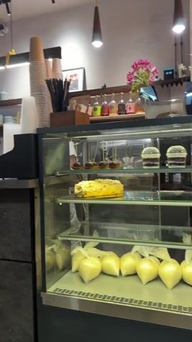 The video pans across a cafe counter. On the counter, there are stacks of paper cups, a black paper cup holder, a tablet device, and a yellow credit card reader. Behind the counter, there are shelves with mugs, bottles of colorful liquids, and a small bouquet of pink flowers. To the right, a glass display case shows pastries on the top shelf and what appear to be bags of cream on the bottom shelf. The counter itself is made of a dark, textured material. In the background, there are exposed brick walls, a green curtain, and hanging pendant lights. A chair is visible in the lower left corner.