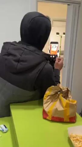 From a medium shot and rear perspective, an individual wearing a hooded jacket is filming inside a room. They are holding a smartphone on a tripod and focusing the camera on a can of Tic Tac mints positioned on a tripod inside a room across from them. On a green table in the foreground are a McDonald's bag and fries.