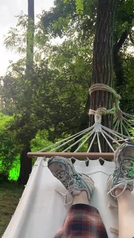 A high-angle outdoor shot shows a person's legs and feet resting on a white fabric hammock in a park. The person wears sneakers and a plaid skirt. The hammock is tied to a large tree on the right side of the frame, and a vast expanse of green trees and bushes is visible in the background under a bright sky. The person's left foot is slightly raised, and the right foot is tucked beneath it. The video is still with a slight breeze rustling the leaves.