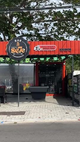 The video shows the exterior of a red shipping container transformed into a bar and convenience store named "BAR SE7E". A black sign with "BAR PARADA SE7E CONVENIENCIA" is visible. There is a delivery phone number "99225.7941" displayed on a white oval sign. Small Brazilian flags are strung across the top of the entrance. A black planter is in front of the store. The ground is paved with bricks. The street is visible in the foreground. Trees and sky are in the background.