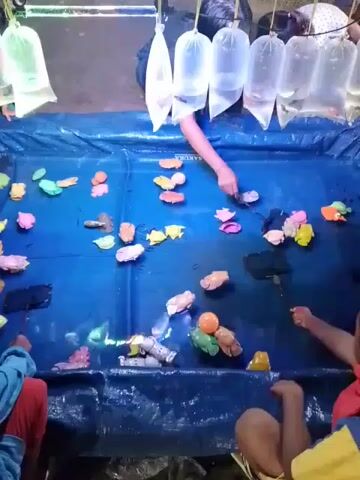 The scene displays a playful game where several children are engaged in fishing for colorful plastic toys from a makeshift pool. The pool, lined with a blue plastic sheet, is filled with an assortment of toy fish and other aquatic creatures, all in vibrant colors. Suspended above the pool are several plastic bags, each containing a live goldfish, presumably the prize for successfully catching a toy. Two of the players are visible, one a young boy in a blue shirt and red leggings who is intently focused on the game, seated on a blue stool. Another individual, possibly an adult, is reaching into the pool with a small net, assisting or overseeing the game. The environment is simple, with an emphasis on the colorful display of toys and the anticipation of winning a goldfish.