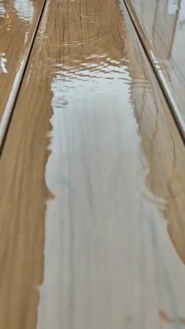The video shows a close-up view of wooden planks with water on them. Raindrops are creating ripples on the surface of the water. The lighting is soft and diffused, suggesting an overcast day or indoor lighting. The wood appears to be smooth and possibly varnished, reflecting the water and light. The camera is positioned low, looking down the length of the planks. There are no people or other objects visible in the frame. The overall impression is one of a peaceful, perhaps melancholic, rainy day scene.