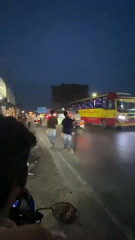 The video displays a night scene on a wet, multi-lane road from the perspective of a person riding a motorcycle. A large bus, painted red, yellow, and blue, travels from right to left across the frame. Multiple pedestrians are visible on the left side of the road, and a motorcyclist is seen on the right. The road surface reflects the bright headlights and taillights of various vehicles. As the first bus moves past, the camera shifts slightly, revealing distant streetlights and oncoming vehicle headlights. Another yellow bus with a red stripe then approaches from the distance, its headlights prominently visible. The arm and plaid shirt of the person filming are visible in the foreground throughout the video.
