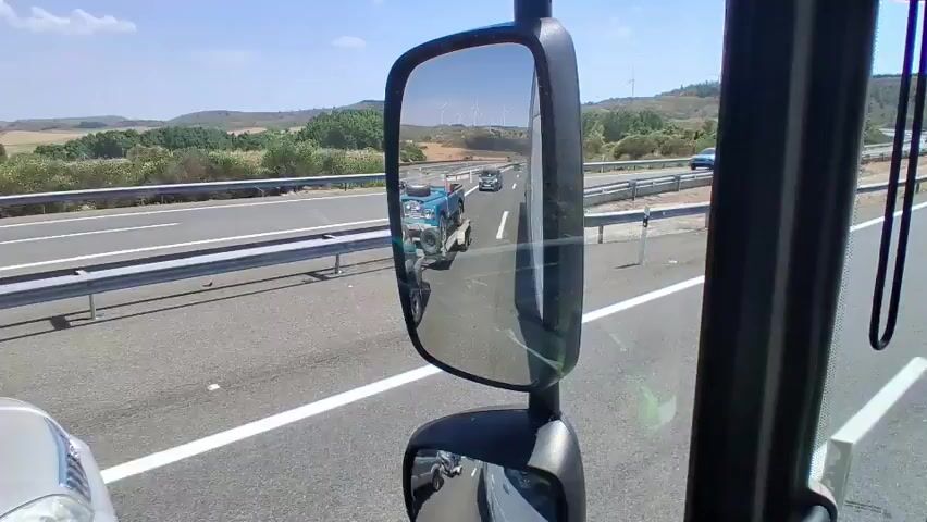 The video captures a perspective from inside a vehicle, likely a truck or bus, driving on a highway. The main focus is the side mirror, which reflects the road behind and to the side. In the mirror, a silver SUV is visible in the right lane, followed by a blue vehicle on a trailer. Further down the road, another vehicle is seen in the distance. The environment appears to be a sunny day with a clear blue sky, and the highway is surrounded by rolling hills and vegetation. The road has multiple lanes and guardrails. The camera also shows a portion of the vehicle's interior, including a black cord hanging down. The video progresses by slightly changing the angle and perspective, showing different vehicles in the mirror's reflection, including a black car and a different silver SUV parked on the side of the road. The video appears to be shot while the vehicle is in motion.
