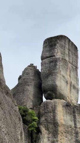 The video shows three rock formations against a cloudy sky. Two of the formations are quite close to each other. On the left rock formation, near its peak, stands a person. The person is holding a white rope or line.