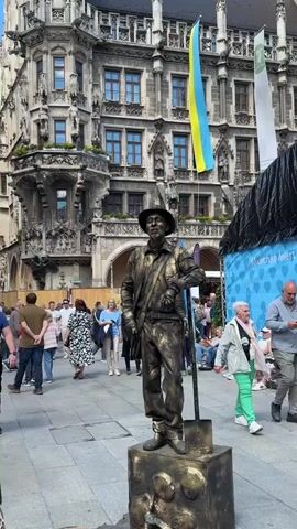 The video shows a street performer covered in gold paint, dressed as a worker, standing on a pedestal. He is holding a shovel and striking a pose, appearing to be a living statue. The performer is in a public square, likely a tourist area, with many people walking around him. In the background, there is a large, ornate building, possibly a historical landmark, and flags are visible. Some people are watching the performer, while others are passing by. The lighting suggests it is daytime.