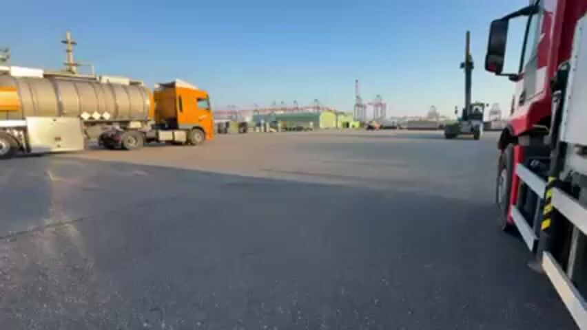 The camera is static, capturing a view of a large industrial area under a clear, blue sky. In the foreground, sections of a red truck are visible on the right, along with a tanker truck with an orange cab and a silver tank driving away on the left. To the left of the tanker truck, there is a complex industrial structure of pipes and towers, suggesting a plant or refinery. In the distance, more equipment and structures can be seen. There is a forklift off to the right. The ground is paved.