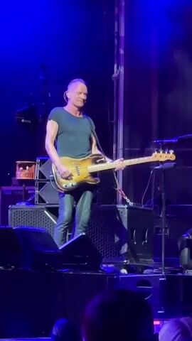 A man playing a bass guitar on a stage. He is wearing a dark t-shirt and dark pants. He has short, gray hair and is looking to the left. The stage is lit with blue lights and there are speakers and other equipment on the stage. There are people in the foreground watching the performance.
