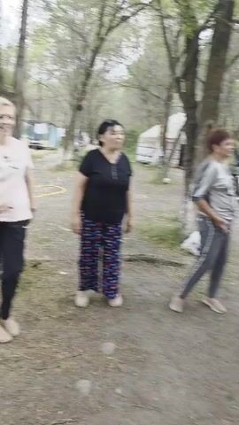 A group of women are outdoors in a wooded area. One woman in a pink shirt and black pants is smiling and dancing. Another woman in a black shirt and patterned pants stands to her right. A third woman in a white shirt and pink jacket with red pants is on the left, moving her arms. A fourth woman wearing a black jacket, white shirt, black pants with stripes, and a headscarf is also visible. In the background, there is a small building with people around it and a yellow hose on the ground. The environment appears to be a park or campground.