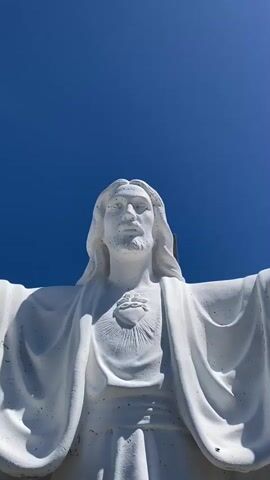 A low-angle, close-up shot features a white statue of Jesus Christ against a clear blue sky. The statue is rendered in a realistic style, with flowing hair and a beard. A heart symbol with rays emanating from it is depicted on the statue's chest, just below the neck. The statue's arms are outstretched to the sides. The texture of the statue shows some imperfections, suggesting it may be made of concrete or a similar material. The bright sunlight creates strong shadows, highlighting the contours of the statue. The top of the statue's head is not visible in the frame, and the bottom of the statue is cropped out.