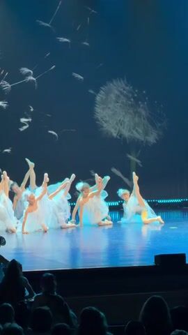 A group of young girls in white dresses perform a synchronized dance on a stage with a large dandelion projection on the background. The girls are mostly in the middle of the stage, some are on their knees with their arms extended to the sides. The stage has a blue and green hue with some light reflections. The audience is visible at the bottom of the frame, watching the performance.
