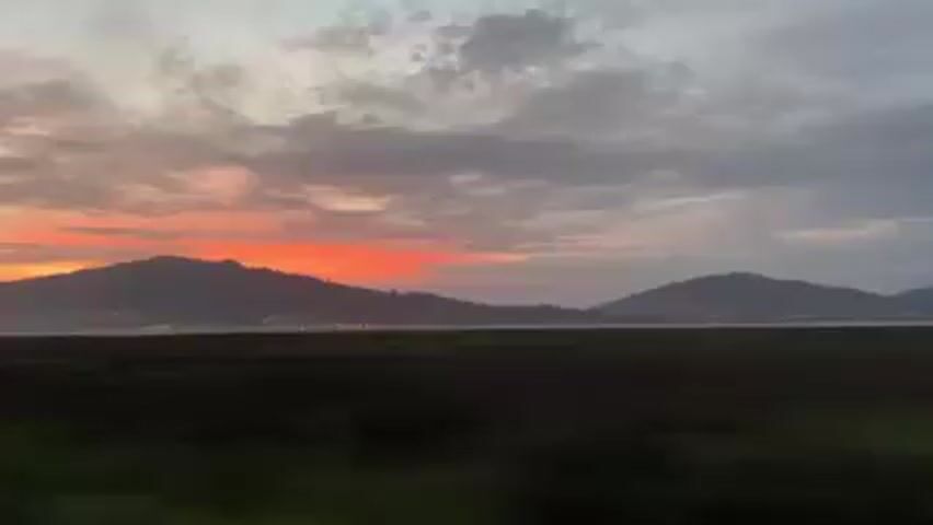 The video shows a landscape at sunset, viewed from inside a moving vehicle.  The sky is a mix of pink, orange, and gray clouds. In the distance, a large hill or small mountain is visible, silhouetted against the bright horizon. A flat, green, grassy area stretches out in the foreground.  A road with a guardrail is present on the near side of the landscape.  The camera is shaking slightly, due to the movement of the vehicle.