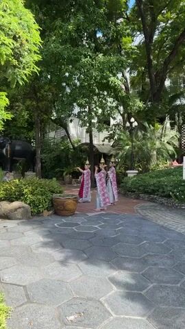 Three women wearing traditional Thai clothing and headwear are performing a synchronized dance in a garden. They are on a stone path with patterned tiles, surrounded by trees, bushes, and plants.  A building with a white facade can be seen in the background. To the left of the dancers is a large decorative pot and a statue of an elephant. A man is sitting to the left of the statue.  The women are smiling as they move their arms and hands in graceful gestures.  The weather appears to be sunny.