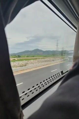 A white truck with a silver cab and a metal cage on its trailer is driving on a highway. The view is through a bus window, with curtains partially obscuring the sides. The truck is moving from left to right across the frame. The environment outside the window includes a road, fields of green grass, hills in the distance, and power lines. The sky is overcast. The camera appears to be moving, as evidenced by the slight blur and the shifting perspective.