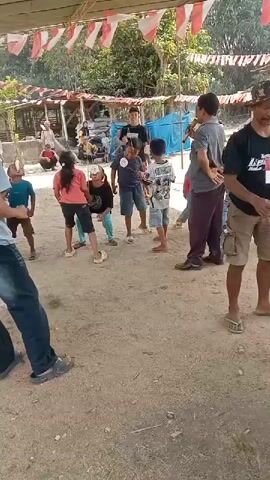 A group of children are participating in a game of tug-of-war outdoors. Red and white flags are strung across the top of the frame, suggesting a festive occasion. Several adults are present, observing the game and possibly acting as organizers or referees. The ground is dirt and sand.
