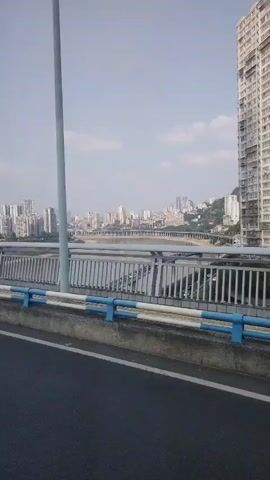 The video shows a view from a bridge in a city. The bridge has blue and white railings.  In the distance, there is a river and a cityscape consisting of many high-rise buildings. The sky is overcast and hazy.  The video appears to be taken from a moving vehicle.