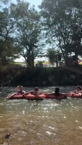 Several people are drifting down a river on inner tubes. They are connected, forming a line. There are trees along the river bank. Behind the trees, a building is partially visible. The weather is sunny.