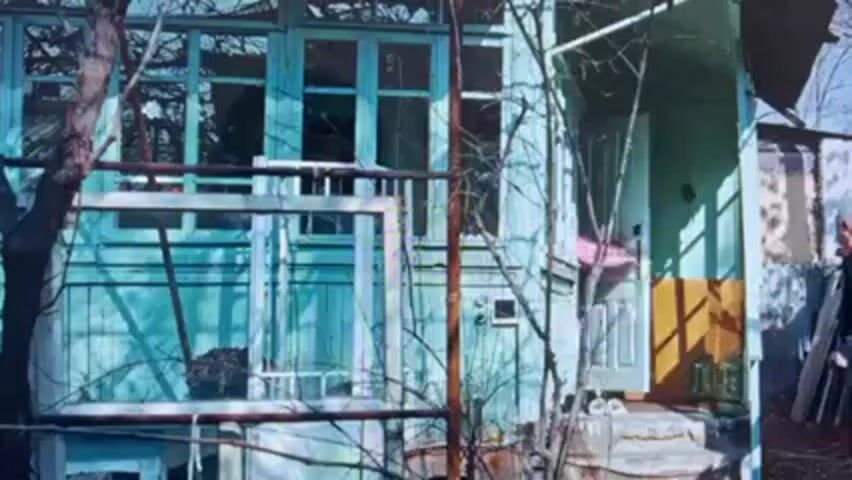 A young woman in a pink sweater and blue headscarf is attacked with a broken green bottle by an older man with a white beard. The attack occurs outside a rundown, turquoise-colored house. The woman is standing near the front porch, and the man is seen both inspecting the broken bottle and in the process of attacking her. The environment appears to be in disrepair, with exposed brick and other debris visible.