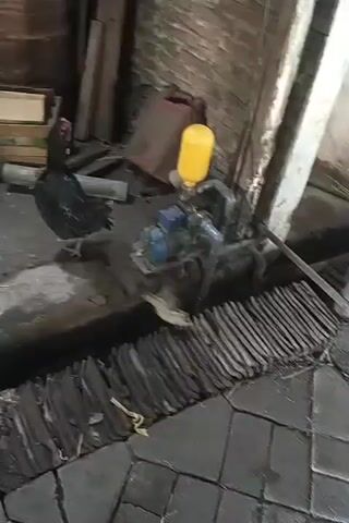 The video shows several chickens walking around a dusty, cluttered area. There are several barrels, wood planks, pipes, and a water pump with a yellow container. One chicken is black and the other two are brown. The chickens are pecking at the ground and moving around the objects in the area. The environment appears to be some sort of workshop or storage area.