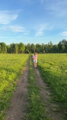 A woman walks on a dirt path through a sun-drenched grassy field in Kerro Massiv, Russia. The sky is bright blue with scattered clouds, and a line of green trees forms the horizon. She is wearing a pink bomber jacket over a striped shirt, light blue denim Bermuda shorts, and pink sneakers. She has long blonde/light brown hair and is holding a smartphone in her left hand. As she approaches the camera, she smiles, appearing relaxed and happy. The golden hour sunlight illuminates the scene, casting a warm glow on the landscape.