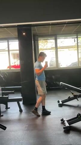 A young man in a light blue t-shirt and gray Nike shorts stands in a gym. He is looking at his phone, possibly taking a picture of himself in the mirror. The gym has black flooring and light-colored walls with large windows showing greenery outside. There are various exercise machines and weight benches in the background.