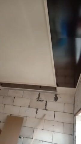 The video shows a room being renovated. A light bulb hangs from the ceiling. The floor is covered in light-colored brick pattern tiles. A white wall borders the tiled floor. A closet or cabinet is partially installed along the wall. There is cardboard on the floor.