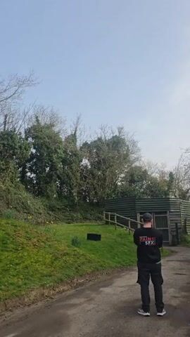A man wearing a black tracksuit stands on a dirt path, facing a grassy area with trees and a small building. The back of his tracksuit has the words "PAINS SFX" in red and white. A black box sits on the grass. The man says "Three, two, one," and a plume of white smoke erupts from the box. The smoke shoots straight up into the sky, then begins to spread out. Another person wearing a pink shirt enters the frame briefly on the right side of the screen. The sky is pale blue with some light clouds.