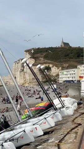 The video shows a view of a pebble beach with several sailboats lined up in the foreground. The sailboats are white with silver masts and rigging, and appear to be small catamarans. Some have numbers on their hulls, such as "1" and "RS CAT 14". In the background, the sea is visible with gentle waves. A cliff face made of white rock with green vegetation on top rises behind the beach. People are scattered on the beach, some sitting, some walking, and some in the water. A building with multiple floors and balconies is situated on the right side of the image, along a paved promenade. Further along the coast, a small church steeple is visible on top of the cliff. Seagulls are flying in the sky. The weather appears to be overcast with patches of blue sky.