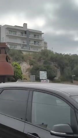 The video shows a large, rusty metal sculpture of a doll-like figure standing on a roundabout. The sculpture has a wide skirt and wears a hat. Behind the sculpture, there is a hill with some greenery and a multi-story building with balconies. A road is visible in the foreground, and a blue circular sign with a white arrow pointing to the right is placed in front of the sculpture. Several other signs are also visible near the sculpture, one with text and an image. The sky is overcast. The camera appears to be mounted on a vehicle moving slowly past the roundabout.
