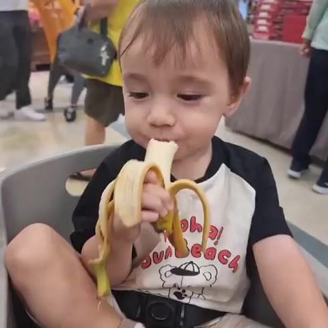 A young child, seated in a shopping cart, eats a banana. The child is wearing a t-shirt with a graphic design. In the background, other shoppers and grocery store aisles are visible. The lighting suggests an indoor environment, likely a supermarket. The video is a continuous, unedited recording of the child's activity.