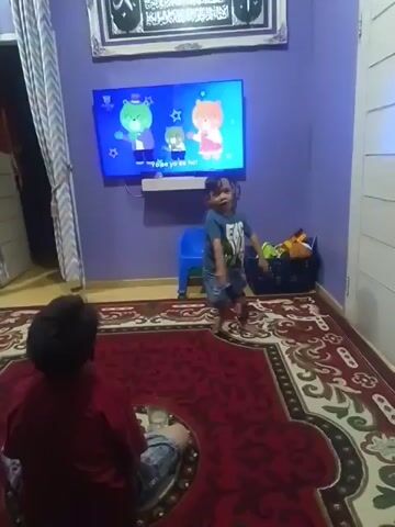The video shows two young children in a room with purple walls and a red rug. A flat-screen television is mounted on the wall and displays a children's cartoon. One child, wearing a blue shirt and shorts, stands and watches the TV, occasionally moving their arms. The other child, wearing a red shirt and jeans, sits on the floor facing away from the camera, also watching the TV. To the right of the TV is a blue plastic stool and a dark basket filled with toys. A window with striped curtains is visible to the left.