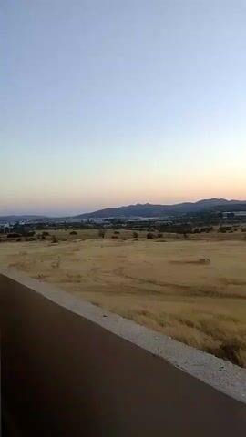 A video captures a landscape view at dusk. The foreground features a concrete structure, possibly a wall or ledge, angled towards the right. Beyond this, a vast, dry, golden field stretches out, with indications of harvested areas or fields. In the mid-ground, sparse trees and some structures, possibly greenhouses or agricultural buildings, are visible. The background is dominated by rolling hills and mountains silhouetted against a sky that transitions from a pale blue at the top to soft shades of pink and orange near the horizon, suggesting the setting sun. The overall impression is one of a rural, arid environment at the end of the day.