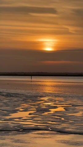 The video shows a sunset over a beach at low tide. The sky is orange and gray. The sun is partially obscured by clouds. The beach is wet and sandy, with pools of water reflecting the sunlight. A single pole is visible in the water.