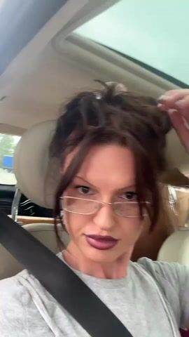 A woman with dark hair pulled up in a messy bun and bangs, wearing glasses and a gray t-shirt, is in the passenger seat of a car. She is touching her hair with her right hand. The car has a sunroof that is open. There are boxes and bags in the back seat. The woman is speaking to the camera.