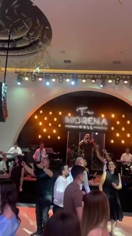 A group of people are dancing at The Morena Hall, with a live band performing on a stage. The band is playing while a man sings with his arms outstretched. The dancers are dressed in both casual and formal attire, and are moving and dancing together on the dance floor. There are other people seated at tables in the background. The Hall appears well lit with a large screen on the wall. There is a decorative hanging piece above the dance floor. The camera moves around, showing various angles of the scene.