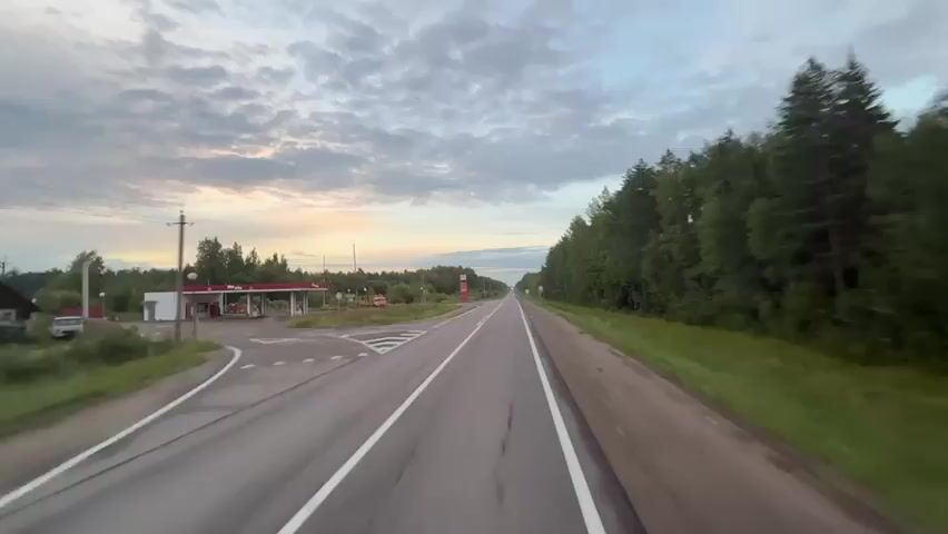 The video shows a first-person perspective of driving on a road. The road is paved and has white lane markings. On the left side of the road, there is a gas station with a red roof and pumps. There is also a red price sign with numbers on it. A truck is parked at the gas station. On the right side of the road, there are trees and a yellow sign with a figure on it. The sky is cloudy with some colors from the sunset. The road stretches into the distance and is lined with trees on both sides. The video captures the movement of the vehicle and the changing scenery.