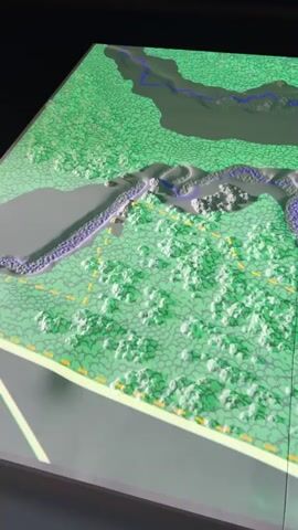 The video displays a 3D topographical model of a landscape. The model features a body of water depicted in blue and purple hues, surrounded by green textured areas representing vegetation or rough terrain. A winding path or road system, marked with dashed yellow lines and solid gray lines, is visible throughout the landscape. On the right side of the model, there are structured areas with grid-like patterns, possibly representing buildings or urban development. Further into the model, on the left side, a section of the landscape is elevated and shaped to represent three standing figures, colored green, possibly sculptures or statues. At the bottom right of the model, there is a structure with red rectangular elements and a large curved roof or canopy, likely representing a building or a significant landmark. The entire model is illuminated by overhead lights, casting shadows and highlighting the three-dimensional relief of the terrain. The video is filmed from an elevated perspective, looking down and across the model.