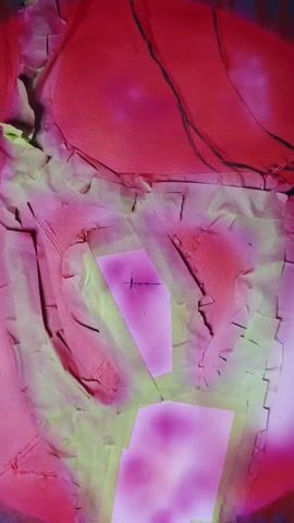 The video shows a close-up of a painting. The painting is abstract, with bold colors and textures. The background is a dark gray, with a large red circle in the center that fades to pink. In front of the circle is a hand-like shape, outlined in bright green spray paint and textured with torn paper. The hand has several rectangular shapes of pink spray paint on it, some of which appear to be blurred or pixelated. The lighting is somewhat harsh, casting shadows and highlighting the textures of the paint.