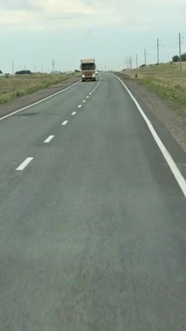 A wide-angle, eye-level shot captures a paved road cutting through a dry, grassy landscape under an overcast sky. The road stretches into the distance, marked by white dashed lines and a solid white line along the right edge. A golden semi-truck with a white cargo trailer is the prominent object in the foreground, traveling away from the viewer in the right lane. Its headlights are on, and the Mercedes-Benz logo is visible on the grille. Further down the road, a white car is also visible, traveling in the same direction. Utility poles line the sides of the road, with some electrical lines extending across the frame. Yellow and black striped markers are placed along the roadside. The landscape appears arid, with sparse vegetation. The overall impression is that of a long drive on a highway in a rural, possibly semi-desert environment. The time of day appears to be daytime, with the cloudy sky suggesting a diffused light. The location context indicates this scene is in Beskaragay rural district, Kazakhstan. There is no discernible text within the video itself, other than what might be on the truck's license plate, which is not legible.